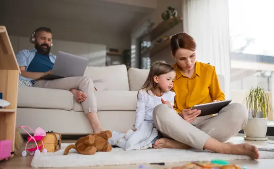 Mutter und Tochter sitzen vor Sofa mit Tablett, der Vater im Hintergrund