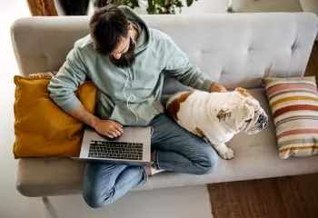 Mann sitzt mit Laptop am Sofa und streichelt eine Bulldogge
